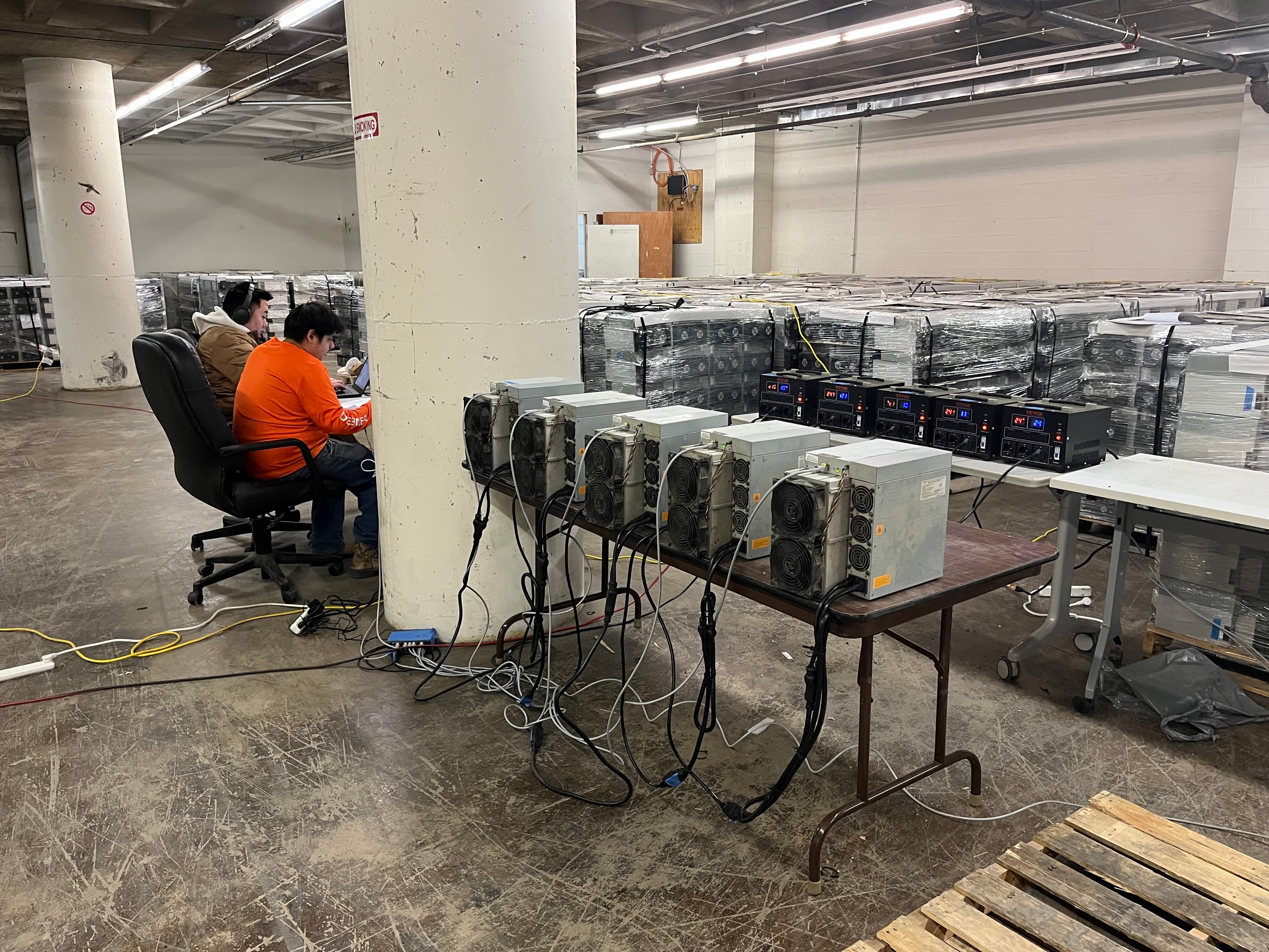 Warehouse staging with ASIC miners on a folding table and stacked equipment boxes